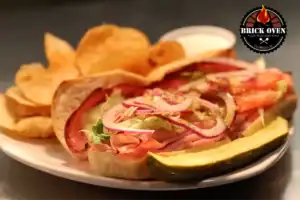 A sandwich with sliced tomato, onions, lettuce, and pickles, served with chips and a side of dipping sauce.